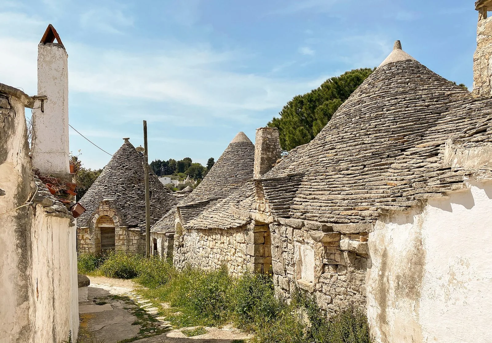Stradina di Alberobello fiancheggiata da trulli in pietra con i tetti conici grigi, muri imbiancati e cielo azzurro