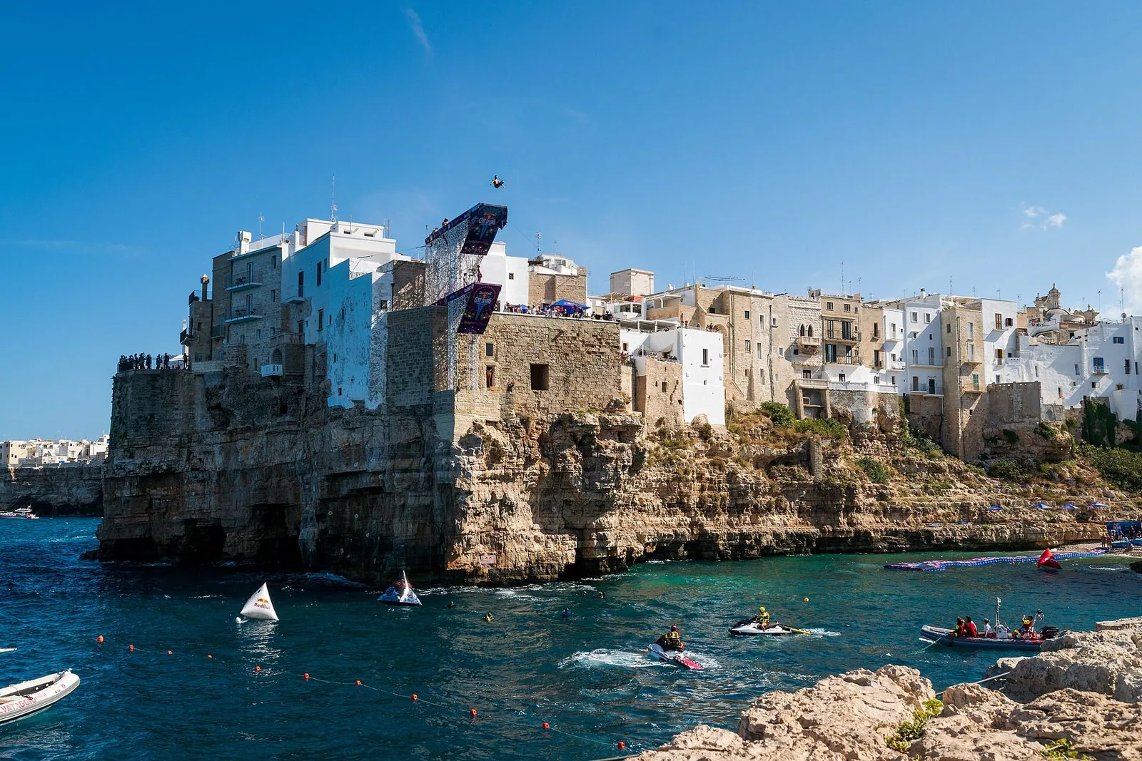 Un tuffatore in volo sopra la piattaforma del Red Bull Cliff Diving montata su un edificio a picco sulla scogliera di Polignano a Mare, con mezzi di assistenza in mare