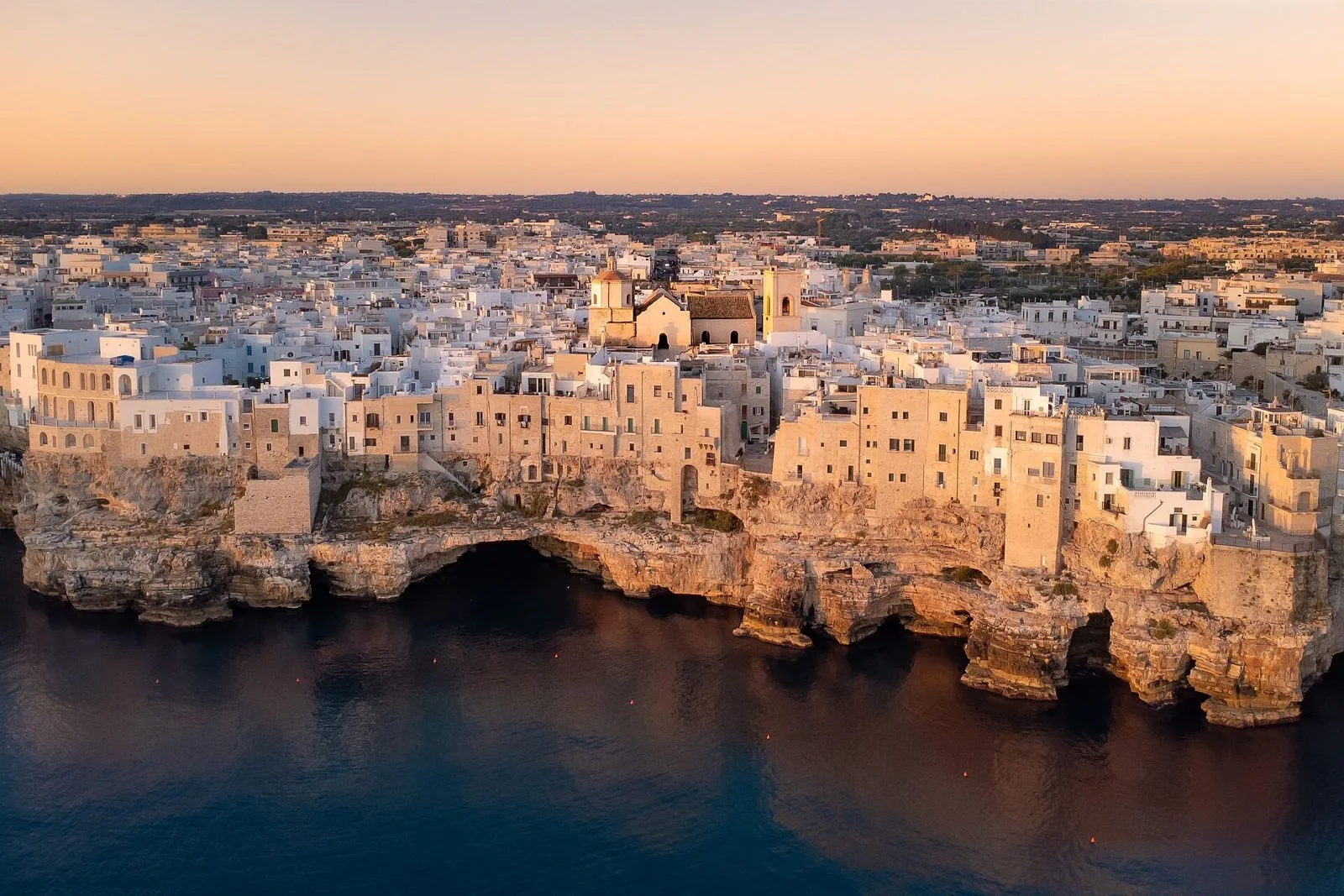 Veduta aerea all'alba del borgo antico di Polignano a Mare, con le case chiare sulla falesia, le grotte alla base e la Chiesa Matrice al centro