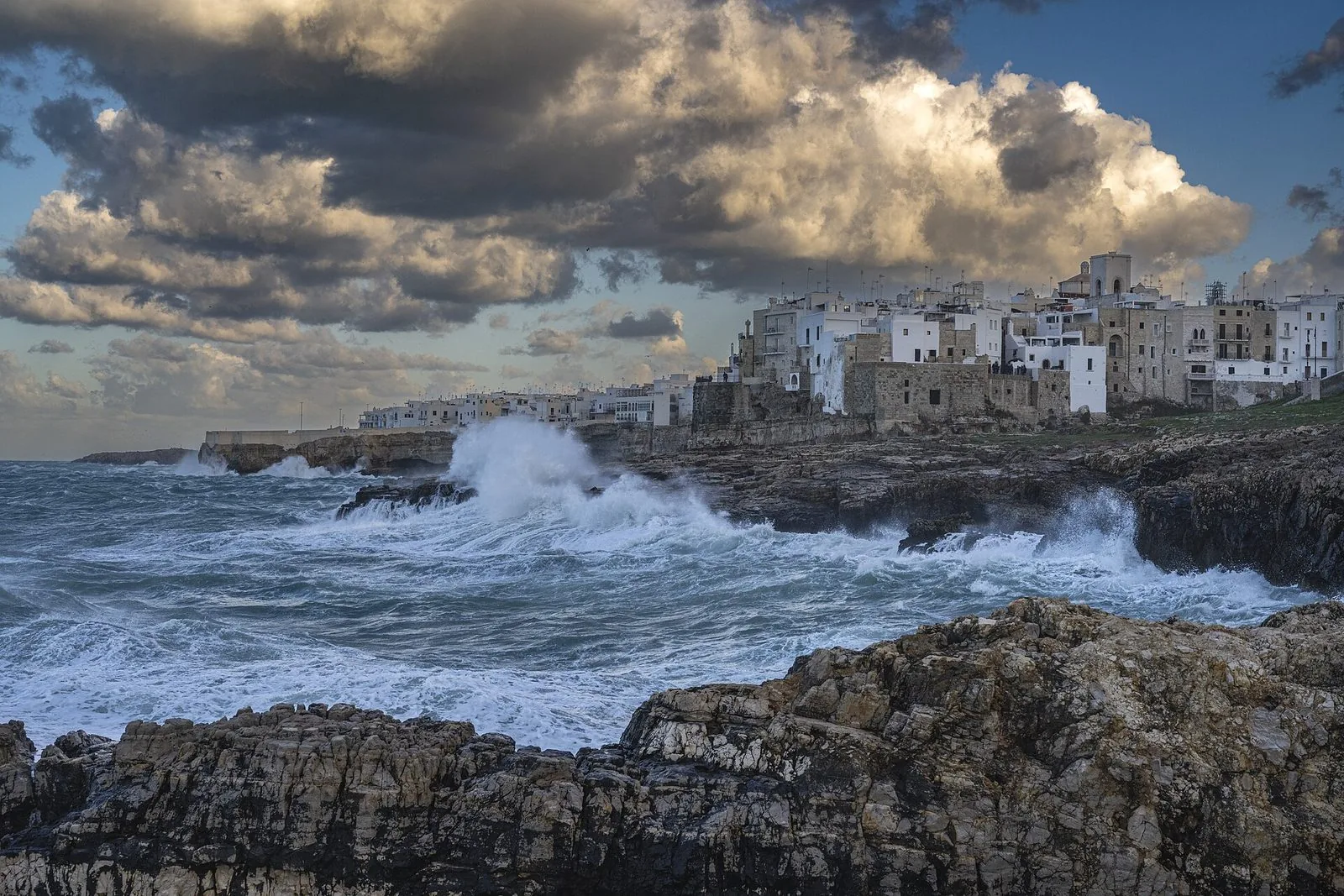 Mareggiata a Polignano a Mare: onde bianche si infrangono sulla scogliera sotto le case del paese, sotto un cielo carico di nuvole