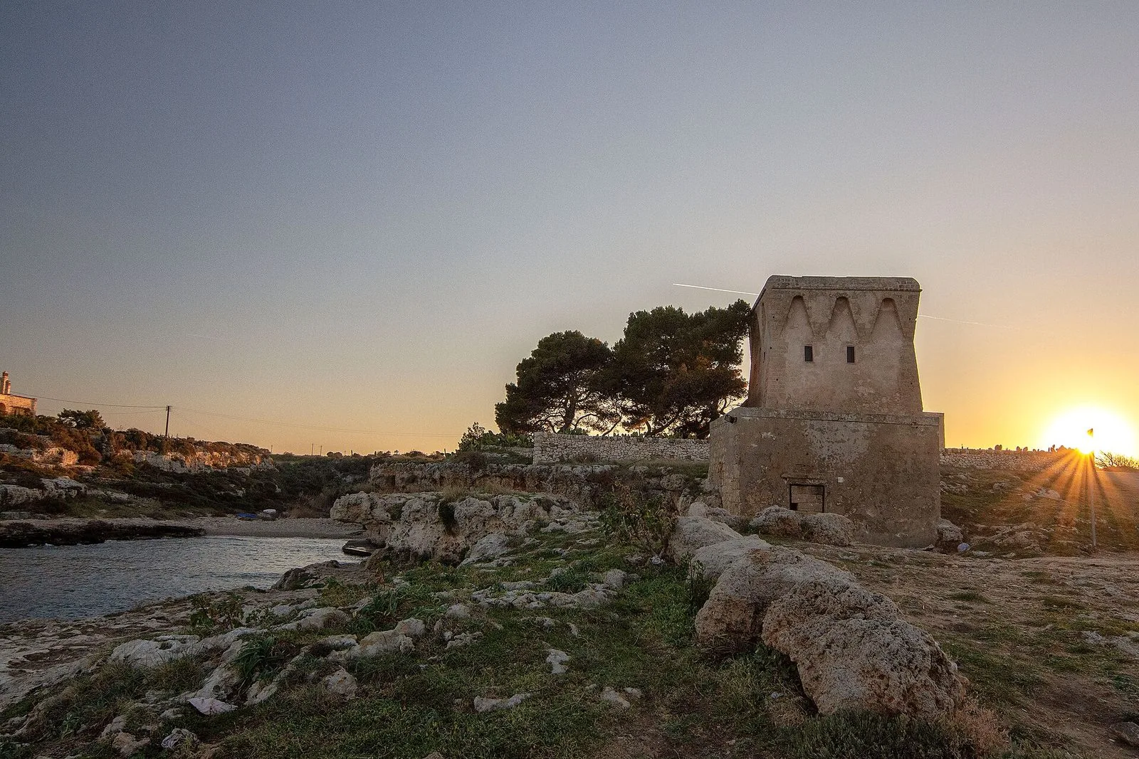 La torre costiera di Cala Incina, sulla costa di Polignano a Mare, al tramonto, con il sole basso all'orizzonte e gli scogli in primo piano